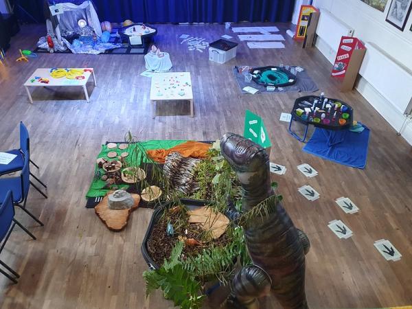 A floor covered in play activities for young children. There is an inflatable dinosaur and paper footprints on the floor next to sandboxes.