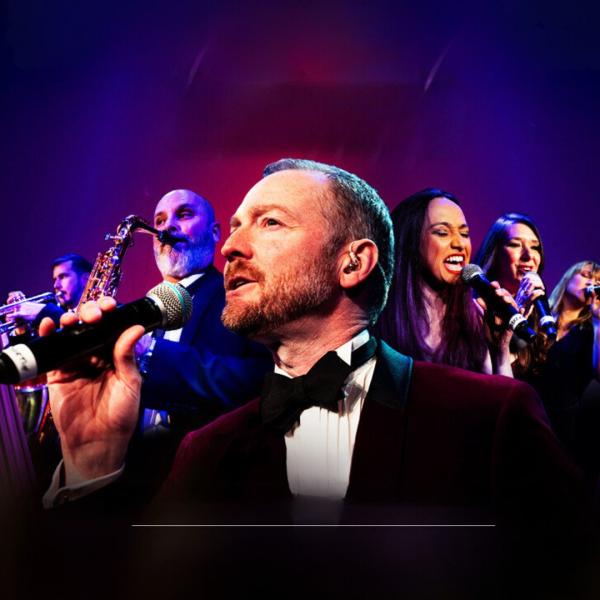 Man in velvet maroon suit and black bow tie holding a microphone to his mouth with musicians who are singing, playing the saxaphone and the trumpet appear in an arrow formation behind him.