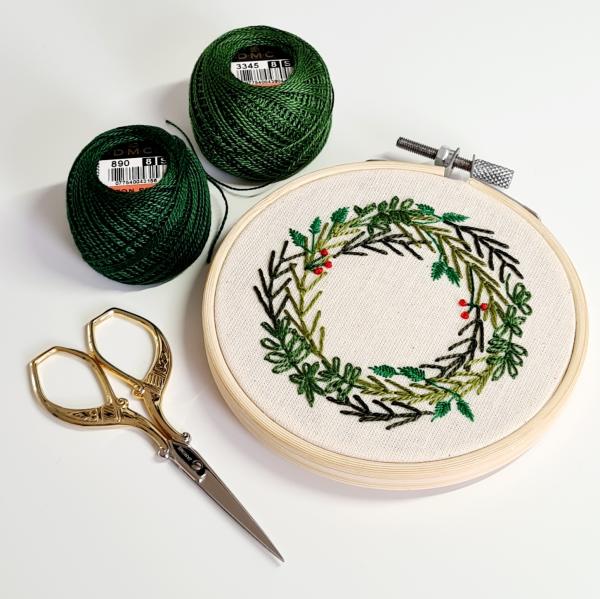 Embroidery hoop with hand stitched wreath in green and brown surrounded by gold pair of scissors and green embroidery thread