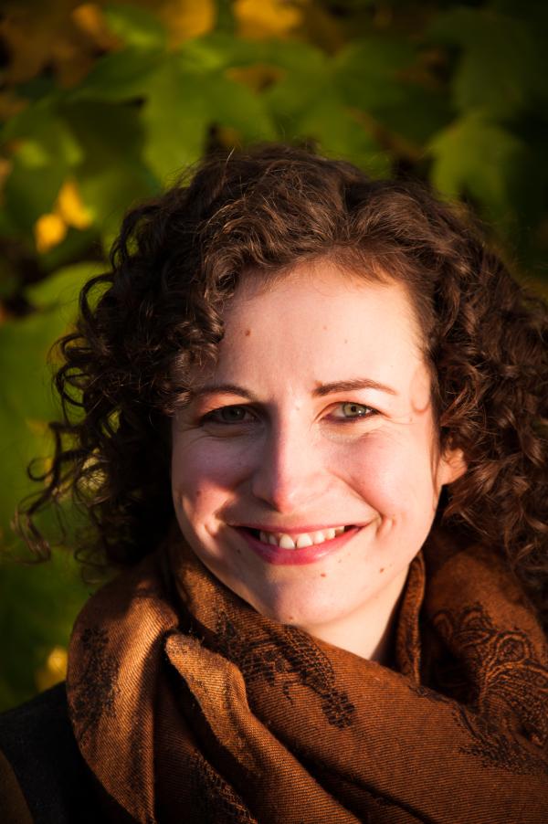 Headshot of a woman in her 30s smiling at the camera with curly brown hair and a brown scarf on.