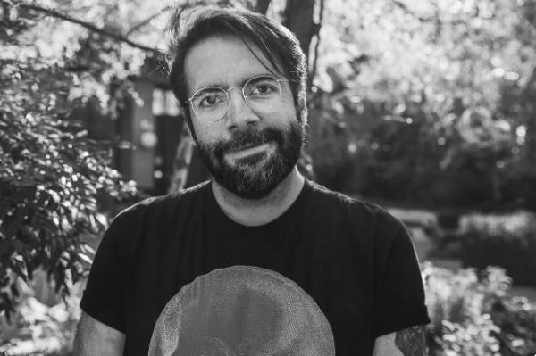 Photo of James Tynion IV, he has short dark hair, a short dark beard and thin metal glasses. The photo is black and white and he's smiling at the camera