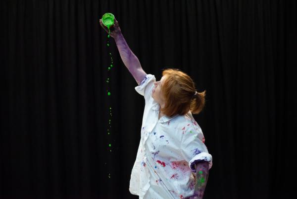 A photo of JoAnne Haines holding au upturned pot of green paint in the air and watching the paint drip out.