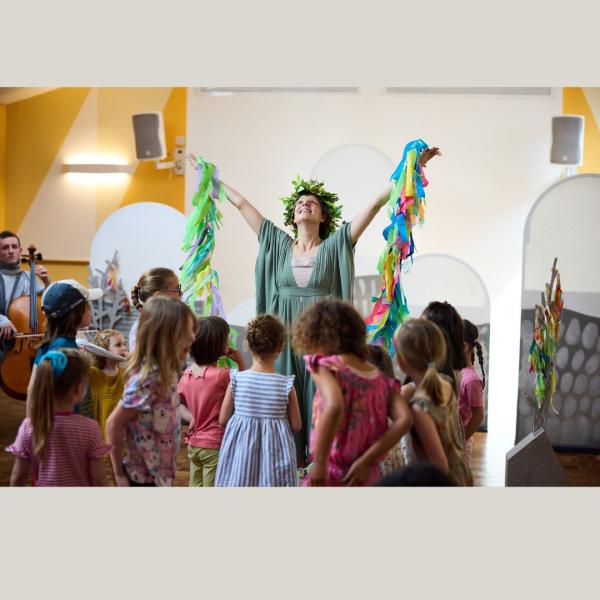 A woman dressed in a green flowing dress, holding up a garland of colourful ribbons, surrounded by children.