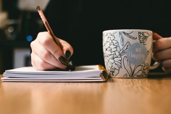 A person with black nail varnish and a black jumper writing in a notebook with a ceramic mug which reads Be Happy