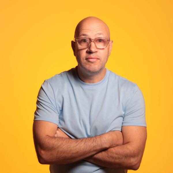 Sam Avery wearing a pale blue tshirt and glasses with his arms crossed against a yellowy-orange background.