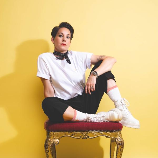 Suzi Ruffell in a white short sleeved shirt, black neck tie with black trousers, white boots and white long socks. Suzi is sat with her feet up and to the side on a fancy red chair with gold legs.