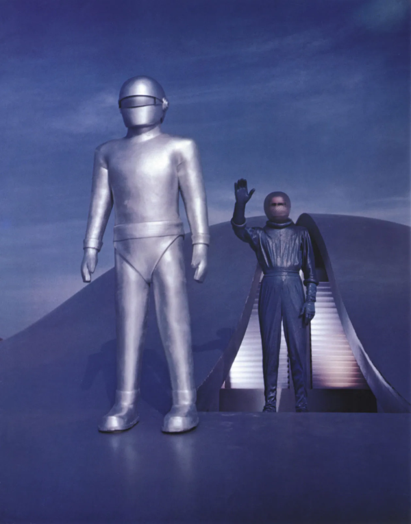 Picture of a large figure in a metal suit and helmet stood next to a smaller figure coming out of a spaceship door.