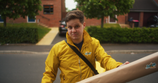 A young man in a bright yellow jacket holds a large parcel towards camera.