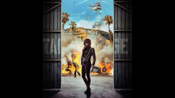 A woman stands wearing a motorcycle helmet, leather jacket and skinny jeans stands in an open warhous doorway with a large explosion behind her.