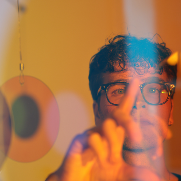 A young man wearing glasses is seen through a number of coloured glass lenses, with blue and orange light over his face. He raises his hand in front of his face to adjust one of the lenses.