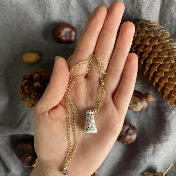 a hand holding a necklace which is a golden chain with a white polymer clay ghost on it which has multi coloured dots as decoration