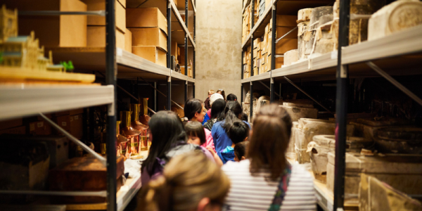 A group of people looking down a narrow corridor with object on tall shelves on each side