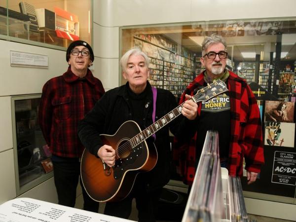 Three men standing in a record store, one holding an acoustic guitar, surrounded by vinyl displays.