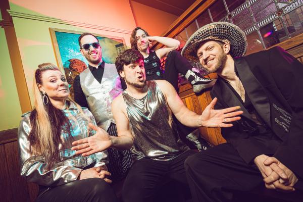 Five band members in colourful, shiny outfits and playful poses, with one in a disco ball hat.