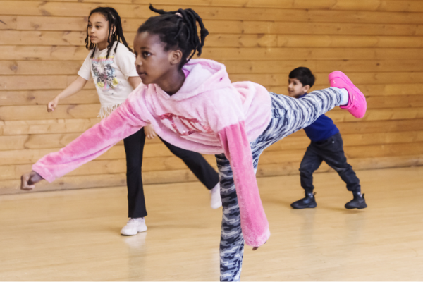 Little girl in a pink jumper leaning forward with one leg extended backwards, two other children dancing behind her 