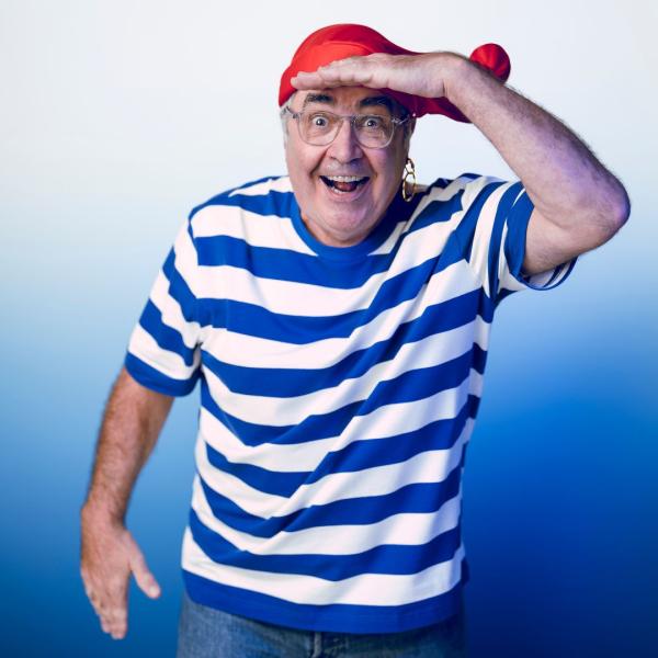 Danny Baker in a blue and white striped t-shirt and red sailor's hat with a gold hoop in his left ear, hand to his forehead as if he's looking for something with a big grin on his face.