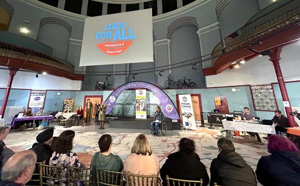 Photo of the 2024 Leeds International Day of Disabled People Marketplace event. at Leeds City Museum. Craig Grimes is speaking into a microphone in front of a seated audience in the main hall where stalls line the edge of the space.