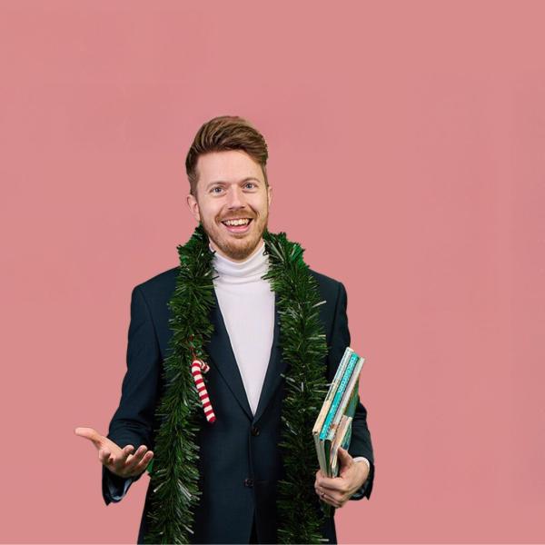 James B Partridge in a navy blazer and white turtle neck with some green tinsel round his neck that has a candy cane attached to it. He is holding some books. A little bit of Christmas tree appears in the top right hand corner.