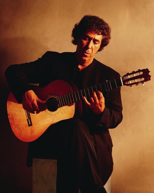 A man sat on a wooden stool playing an acoustic guitar.