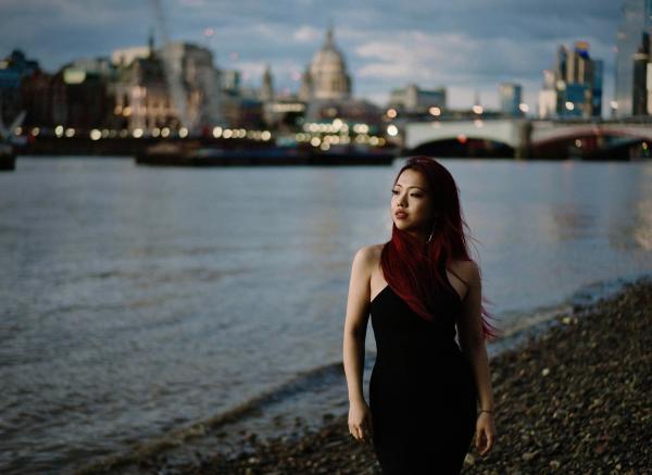 A young Chinese woman standing on the side of the River Thames in London. She has red hair and is wearing a sleeveless black dress.