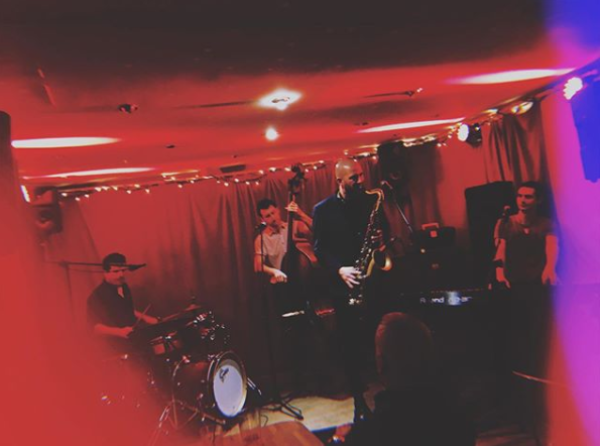 A saxophonist, keyboard player, double bass player and drummer on a red lit stage.