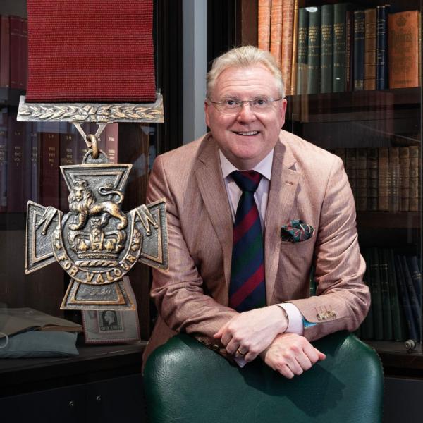 Mark Smith stood leaning on a green leather-backed chair in a library smiling at the camera. A zoomed-in the Victoria Cross appears in the image.