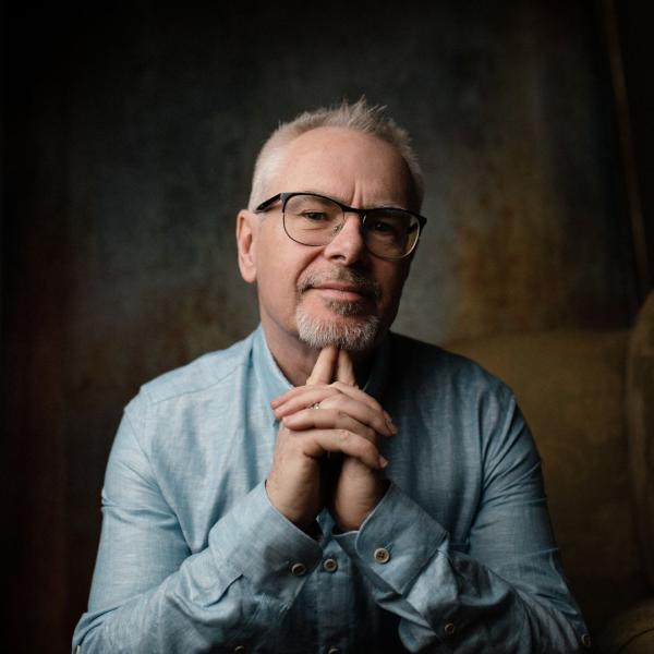 Nik Kershaw wearing glasses in a light denim shirt with his hands together under his chin.