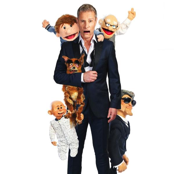 Paul Zerdin in a black suit and undone bowtie with a ventriloquist doll's mouth with other dolls hanging off him including an elderly man, a fox, a baby, a young lad and a man in a black suit and sunglasses.