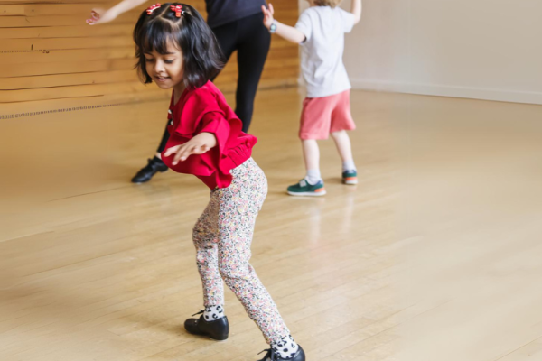 Little girl in red shirt and patterned leggings tap dancing, in the middle of image with a smaller child and adult dancing behind her.