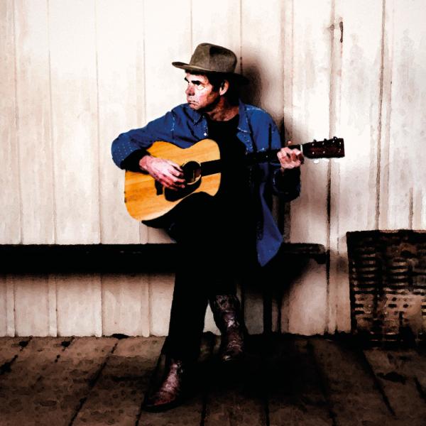 Rich Hall wearing a grey bowler hat playing against an acoustic guitar against a wooden wall.