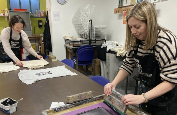 Two women screenprinting images onto t-shirts and tote bags