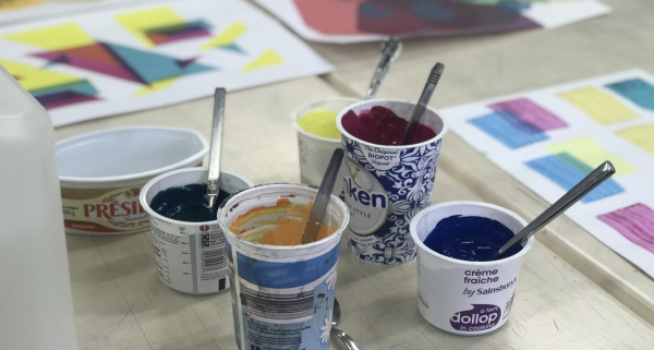 Pots of ink in various colours including orange, blue, red and yellow in front of a selection of screen prints in abstract patterns.