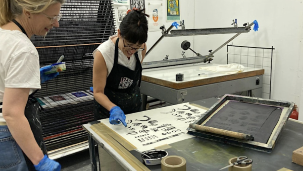 Two women screen printing abstract shapes in black ink onto a white piece of fabric.