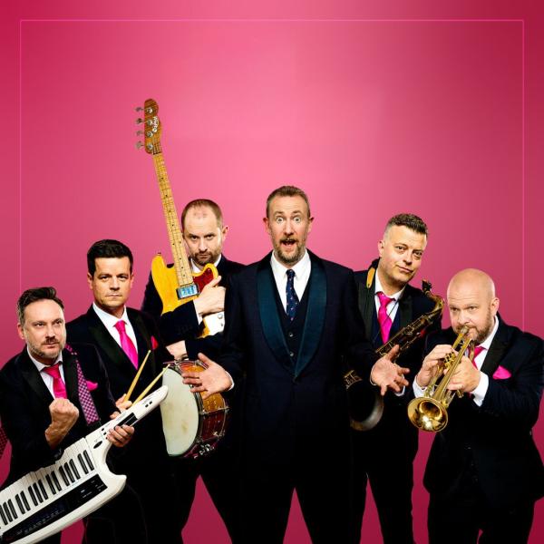 Members of the Horne Section holding their instruments with Alex Horne looking excitedly shocked in the middle