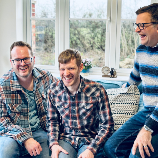 The three members of The Young Uns sat in patterned shirts laughing on a sofa.