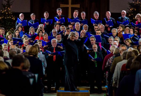 A choir stands between two Christmas trees holding blue folders and singing. The conductor in front of them is turned towards the camera and the audience with one arm raised. The audience has their back to the camera.