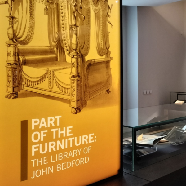 Yellow illuminated exhibition panel which reads "Part of the Furniture: The Library of John Bedford" and has an illustration of a four poster bed, behind which can be seen glass museum cases filled with books.