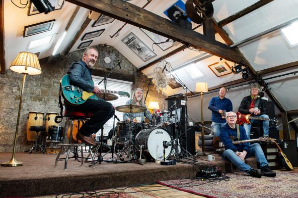 A five-member band in a rustic studio, with guitars, drums, and equipment, surrounded by warm lighting, wooden beams, and framed posters.