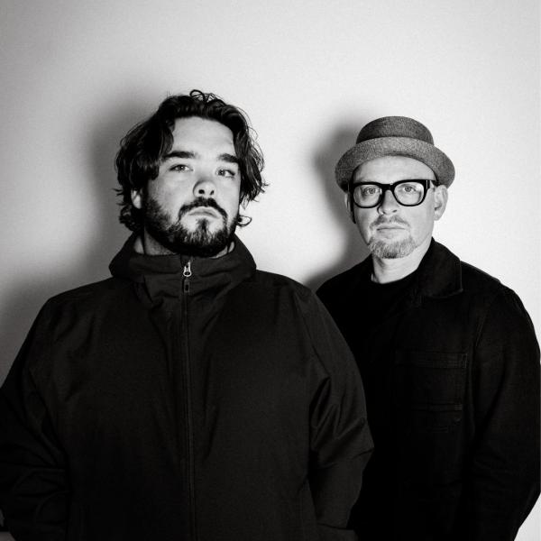 Two men in black and white. The one on the left has shaggy dark hair and a beard and the one on the right is wearing glasses and a bowler hat.