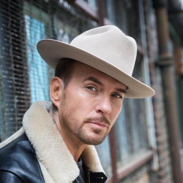 Matt Goss wearing a fur-collared black jacket and beige fedora looking pensively at the camera.