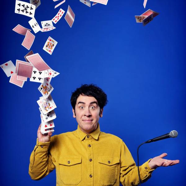 Pete Firman in a mustard-coloured shirt firing a pack of playing cards into the air with his right hand, his left levitating a cord microphone above it.