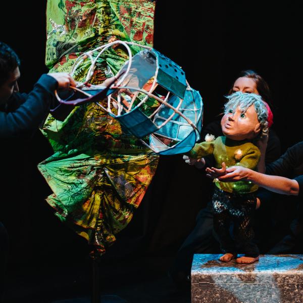 Picture of a barefoot watersprite, with blue eyes, hair poking out under a red cap, clinging onto the blue fish it is riding on. In the darkness, 3 performers hold and operate the puppets, one is gently smiling. The watersprite is looking at a tall weed like fabric that is growing to their side