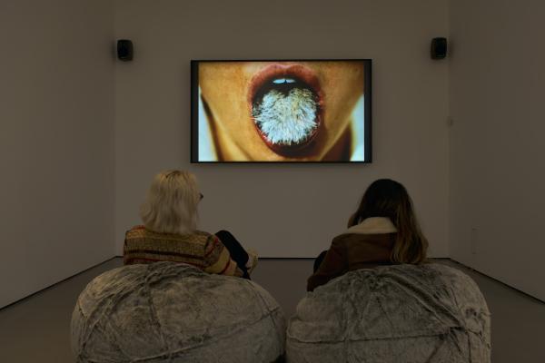 Two people sit on furry beanbags watching a screen showing a woman with her mouth open, sticking out a furry tongue.