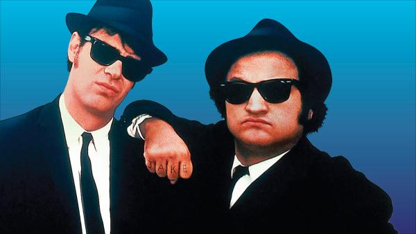 Two men with black sunglasses, suits & hats.