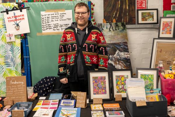 Pyramid's chairperson, Liam, wearing a Christmas jumper in front of a stall selling artwork