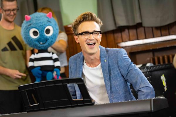 A blonde man with glasses sits at a keyboard, next to a blue monster puppet