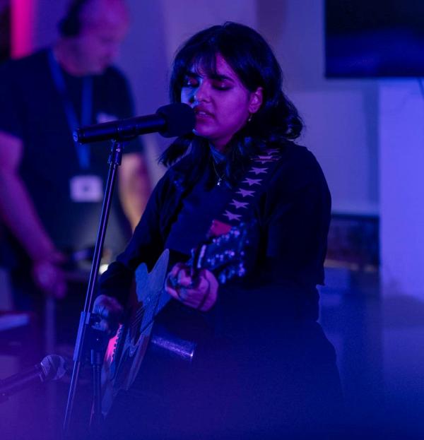 You adult female with dark hair and outfit playing guitar and singing in music library with a blue purple light and person in back ground managing sound levels