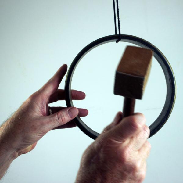 A ring of metal, roughly 30cm in diameter, hanging on a loop of string. A hand is steadying the ring, while a second hand is poised to strike it with a small, square hammer.