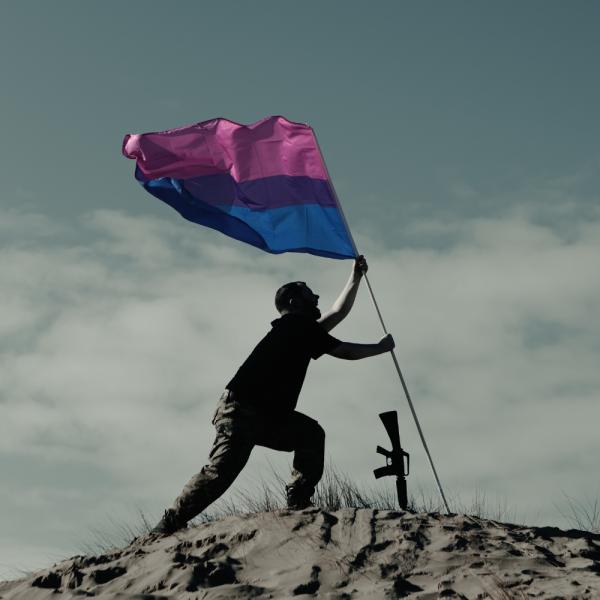 A man at the top of a sand dune sticks a bisexual pride flag into the ground