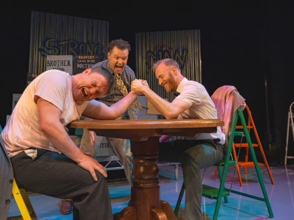 Two men are having an aggressive arm wrestle across a table, while a third man looks on shouting at the match.